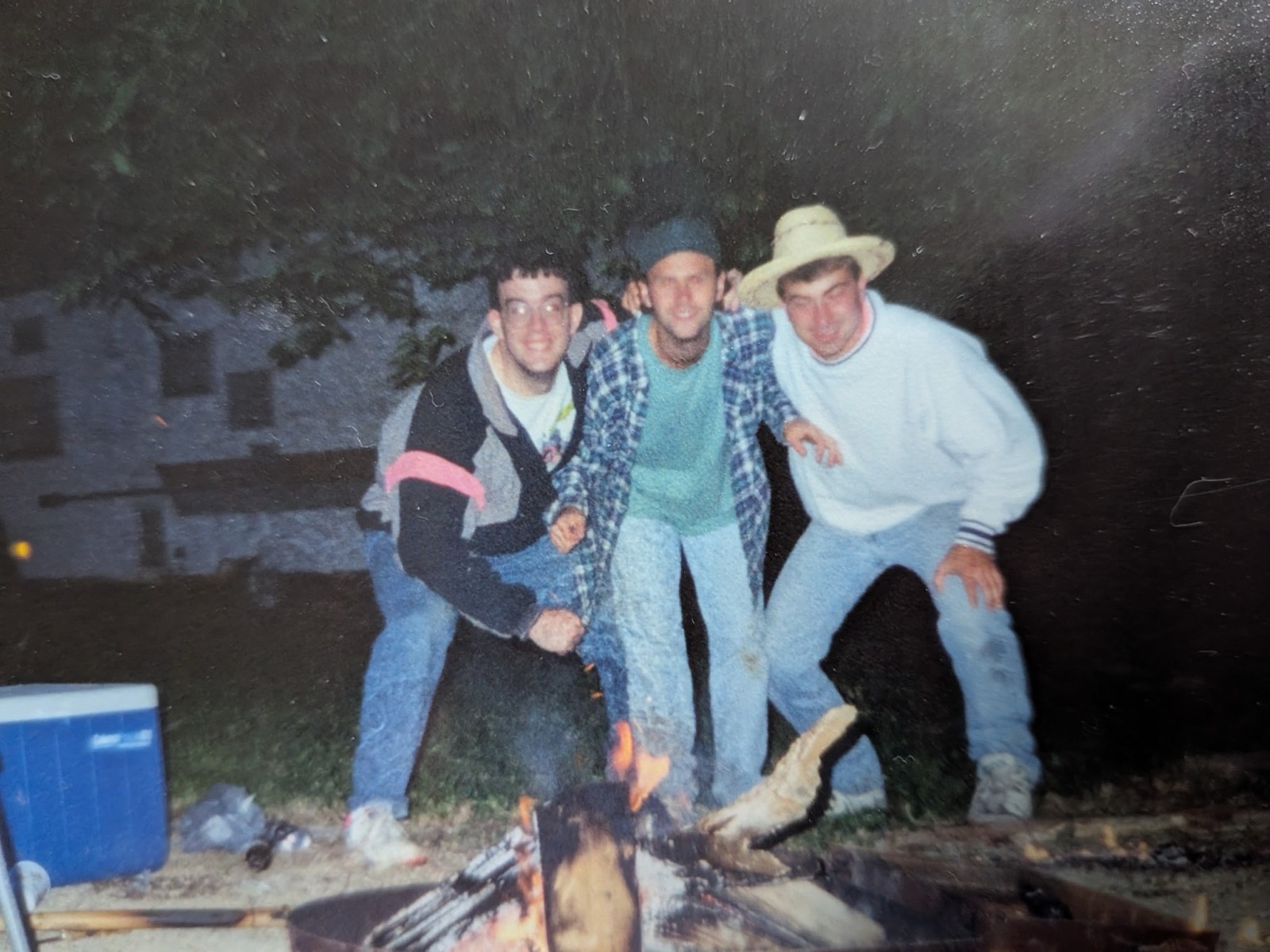 A night flash photograph of three young men leaning in together behind a campfire ring. Left to right: a man in glasses and a black, gray and pink jacket, a man in a teal shirt and open blue plaid flannel with a dark knit cap, and a man in a white sweatshirt and a straw hat. A blue cooler sits at the left edge.