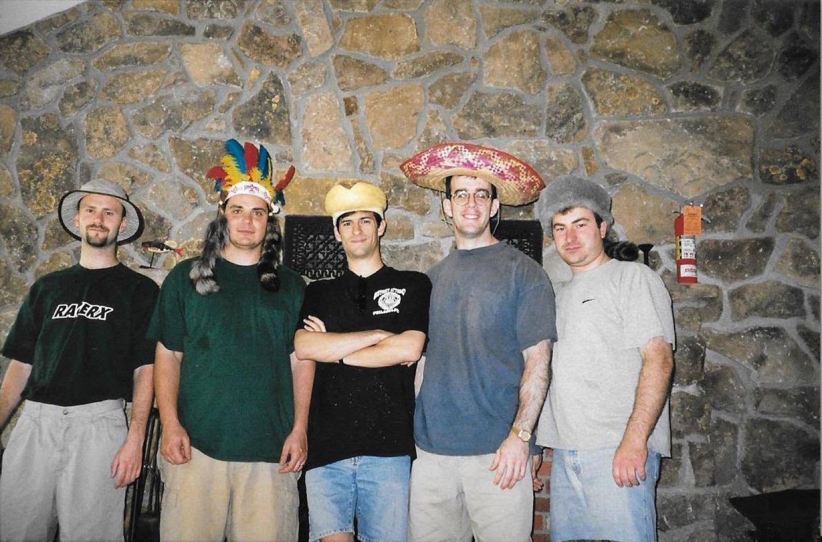 Five young men standing shoulder to shoulder in front of a rough stone wall, each wearing a different silly hat: a wide bush hat with a chin strap, a feathered headdress, a yellow hard hat, a pink straw sombrero, and a fur cap.