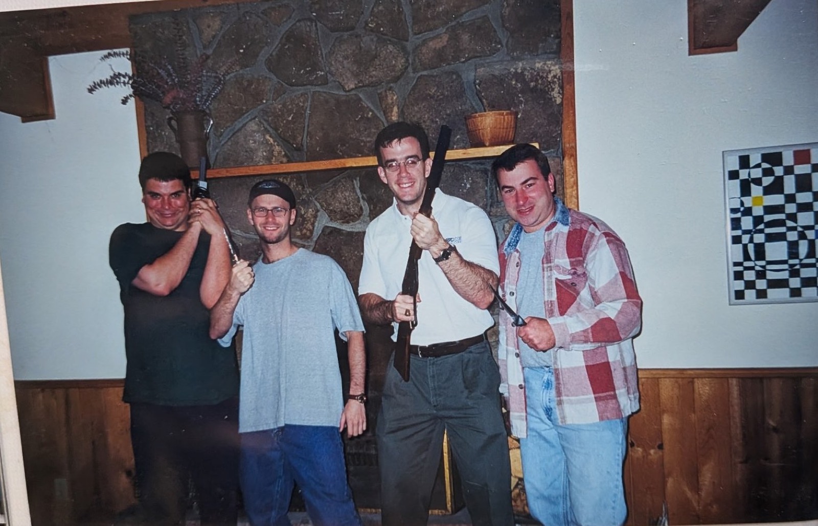 Four men posing in front of a stone fireplace with a wooden mantel in a cabin living room. Left to right: a man in a black shirt holding something upright at his shoulder, a man in a gray T-shirt and dark cap holding a knife, a man in glasses and a white polo holding a rifle upright, and a man in a red and white plaid flannel holding a knife. A black and white checkered painting hangs on the wall at right.