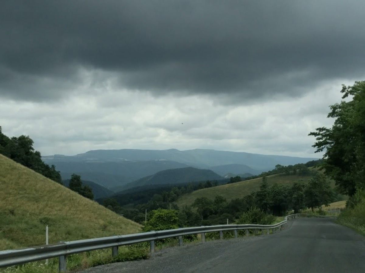A two-lane road curving along a mountainside past a guardrail, layered blue ridges ahead and a heavy dark storm cloud filling the sky above them.