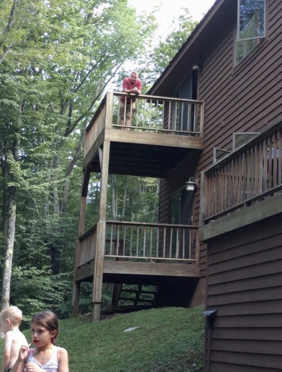 A tall brown wood-sided cabin built into a wooded slope with two stacked deck levels, a man leaning on the upper rail looking down, two children in the grass at the bottom of the frame.