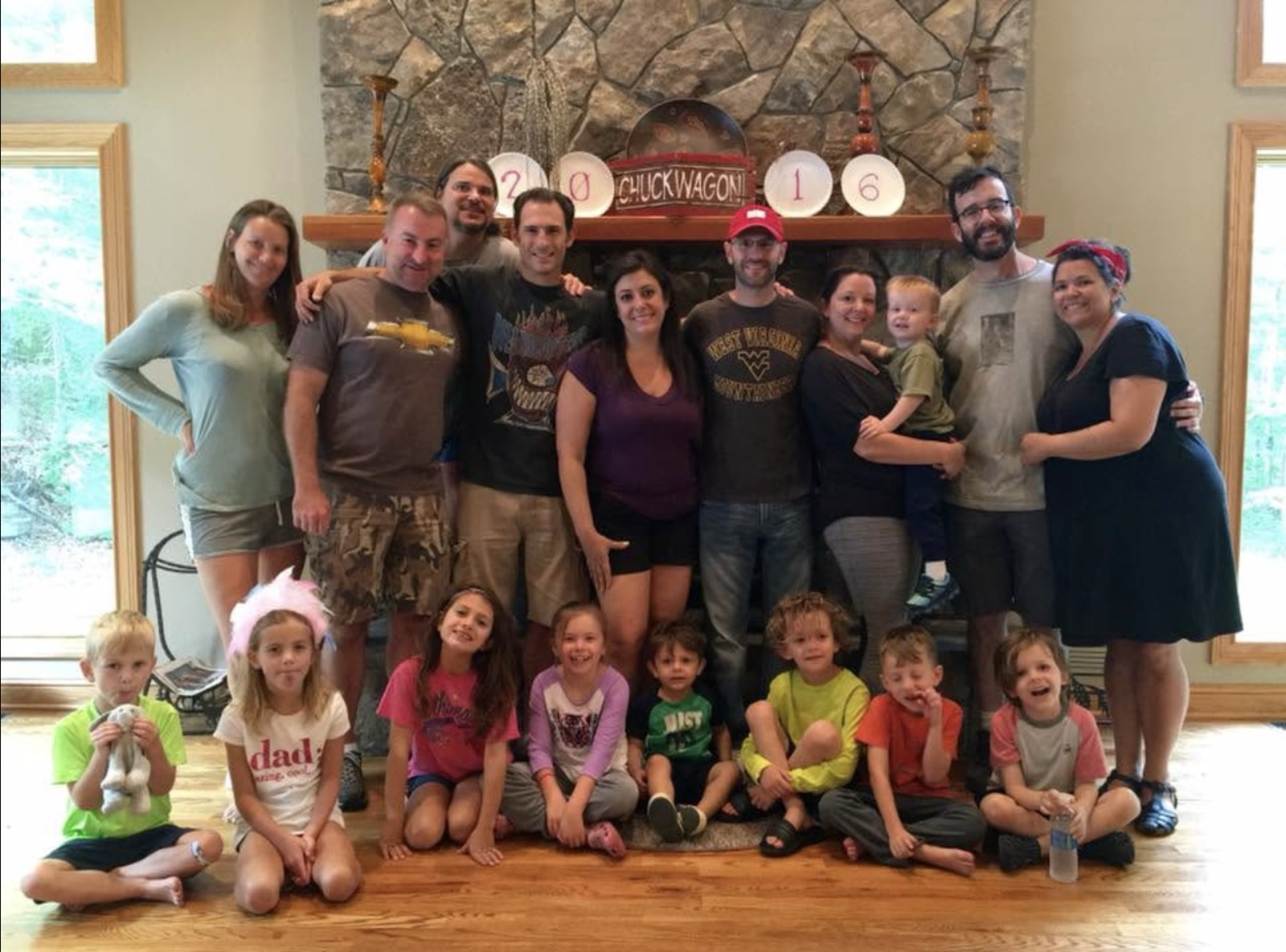 The entire Chuckwagon Gang, adults standing in a row with nine children seated on the floor in front, posed before a tall stone fireplace. Paper plates on the mantel spell 2 0 1 6 around a red sign reading CHUCKWAGON. Left to right the adults are a woman in a pale green top, a man in a brown Chevrolet shirt and camouflage shorts, a long-haired man in gray behind them, a man in a black graphic shirt, a woman in a purple v-neck, a man in a red cap and West Virginia Mountaineers shirt, a woman in black holding a toddler in olive, a bearded man in glasses, and a woman in a navy dress and red headscarf.