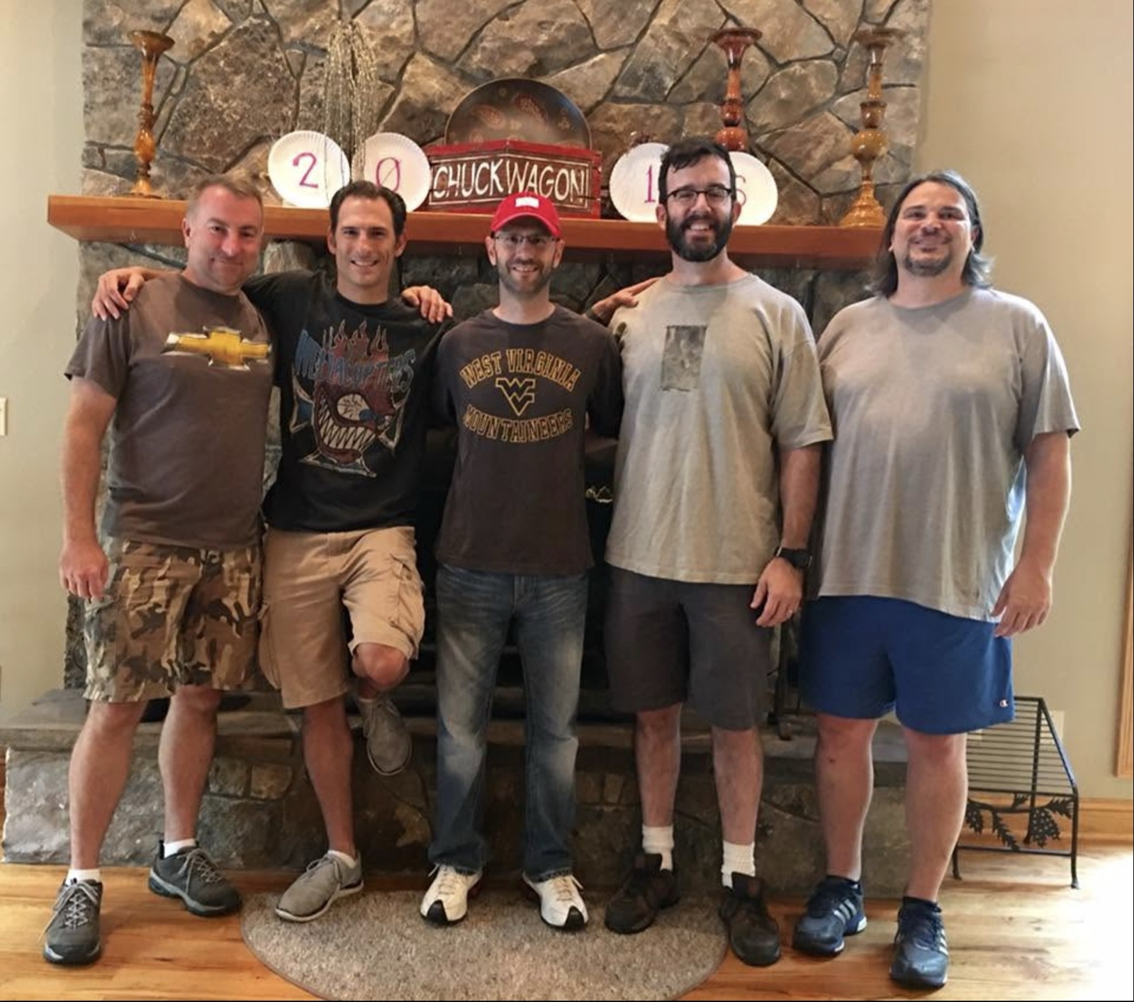 The five founders standing shoulder to shoulder in front of the stone fireplace with the 2016 paper plates and CHUCKWAGON sign on the mantel above them. Left to right: Heath in a brown Chevrolet t-shirt and camo shorts, Brian in a black graphic tee, Shawn in a red ball cap and a West Virginia Mountaineers shirt, Jim bearded in an olive pocket tee, and George in a gray tee with long dark hair.