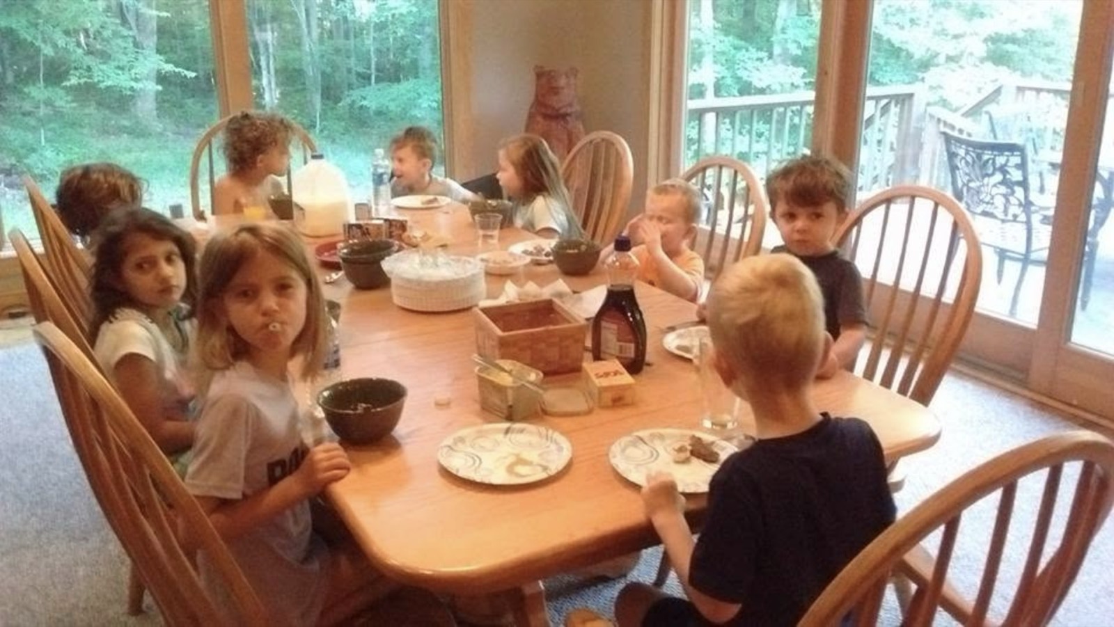 Nine children around a long wooden dining table covered with cereal bowls, a milk jug, syrup and paper plates, tall windows onto green woods behind them and a carved wooden bear standing in the corner.