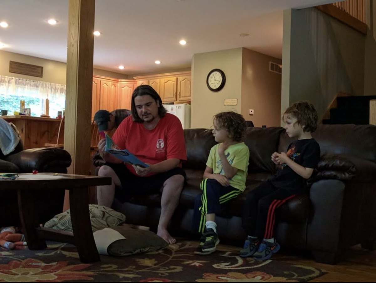 A man in a red t-shirt reading aloud from a picture book on a leather couch while two boys sit along the same couch listening, a cabin kitchen and wall clock behind them.