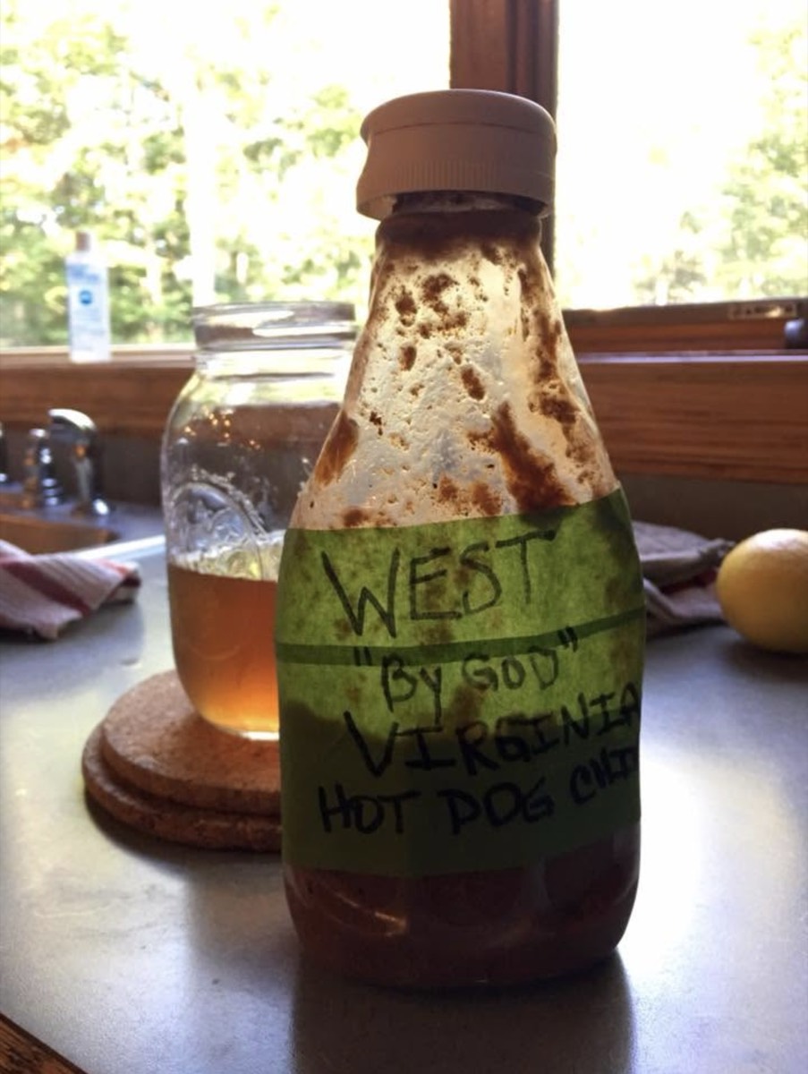 A well-used squeeze bottle on a kitchen counter with a strip of green painter's tape for a label, hand-lettered WEST BY GOD VIRGINIA HOT DOG CHILI, a jar of iced tea behind it.