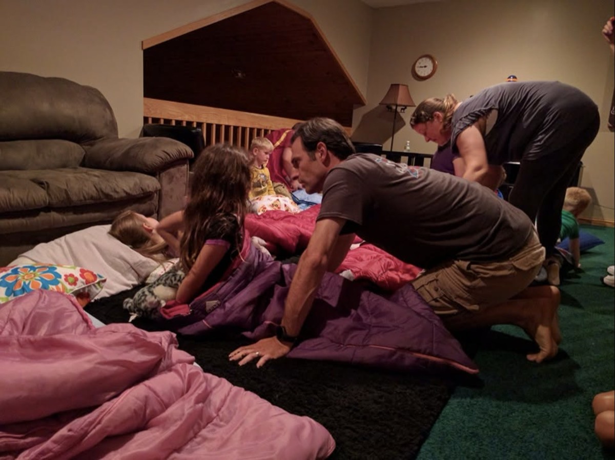 Adults kneeling on a carpeted cabin floor covered in pink and purple sleeping bags and pillows, tucking in several children for the night under lamp light.