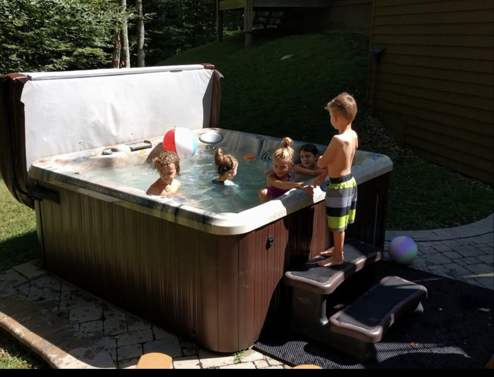 Five children in a hot tub on a cabin patio with the cover propped open behind them, a beach ball floating, one boy standing on the steps about to get in.