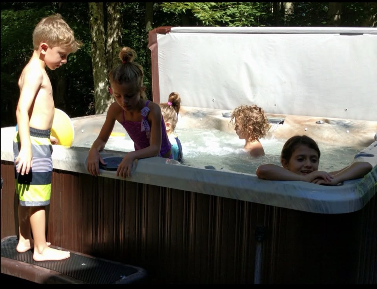 A boy in striped swim trunks standing beside a hot tub looking in, a girl leaning over the edge from inside, three more children in the water behind her.