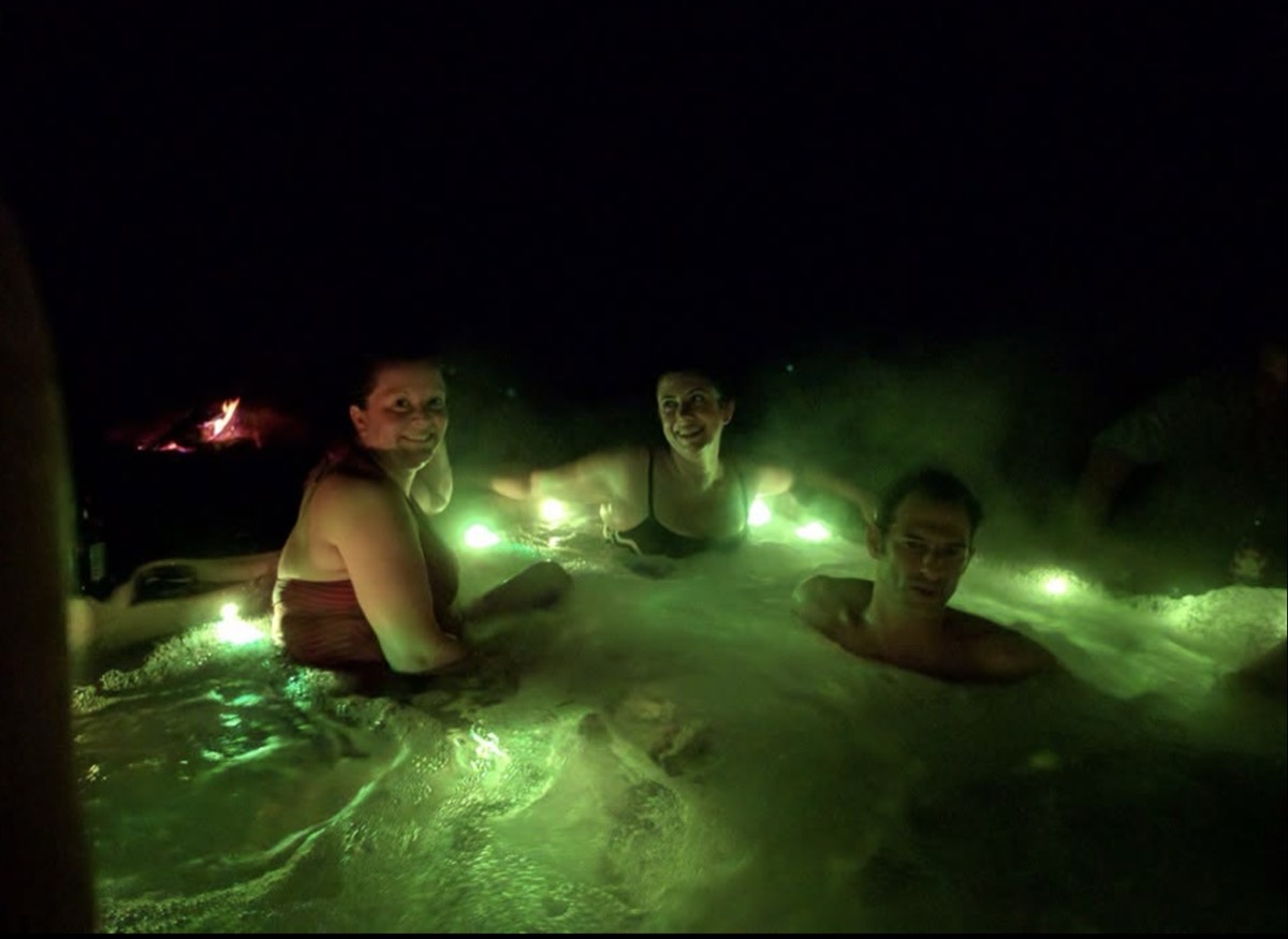 Three adults in a hot tub at night lit green from the underwater lights, steam rising off the water, a small fire burning in the dark behind them.
