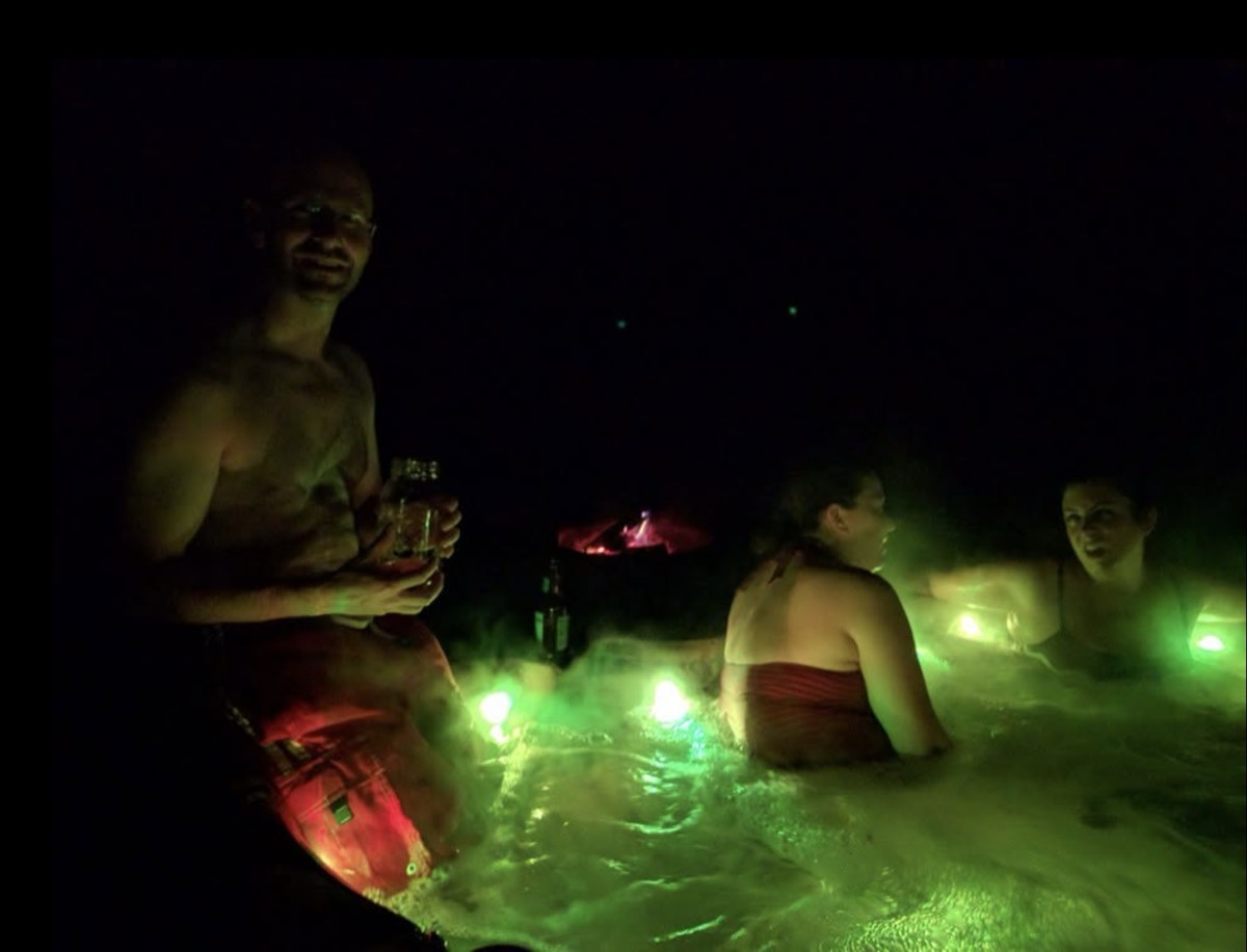 A man standing in a hot tub at night holding a drink, lit green from below, two women seated in the water beside him and a fire glowing in the background.
