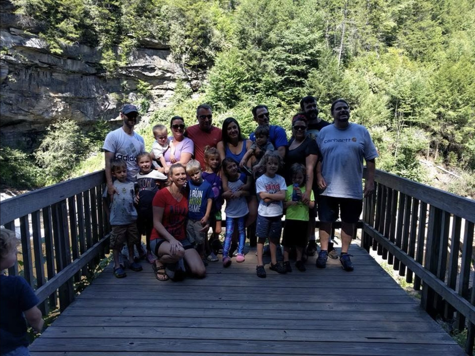The whole gang of adults and children posed together on a wooden boardwalk at Blackwater Falls, a mossy rock face and dense green forest rising behind them.