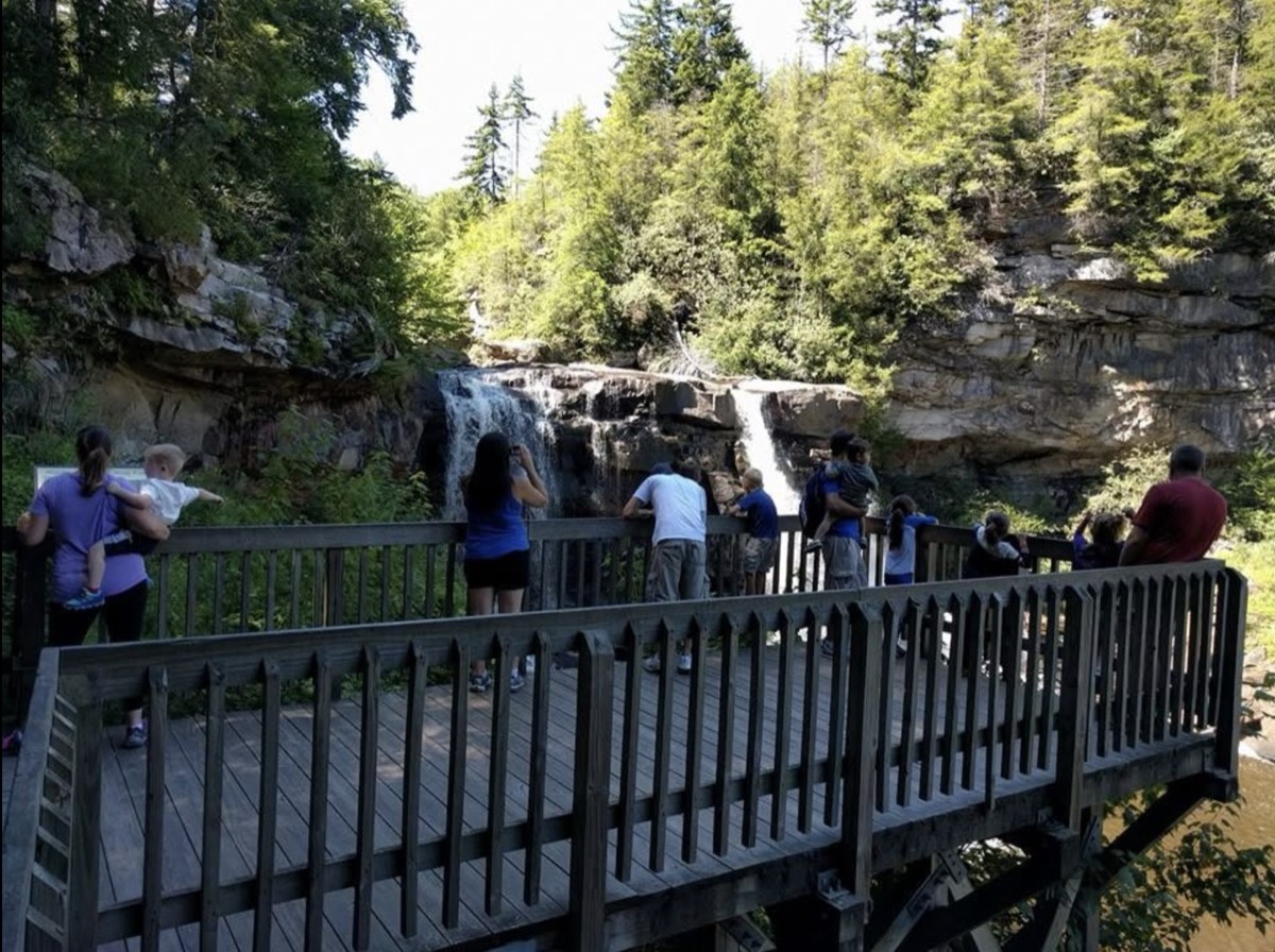 Adults and children spread along a long wooden observation deck above Blackwater Falls, several holding toddlers up to see over the railing.