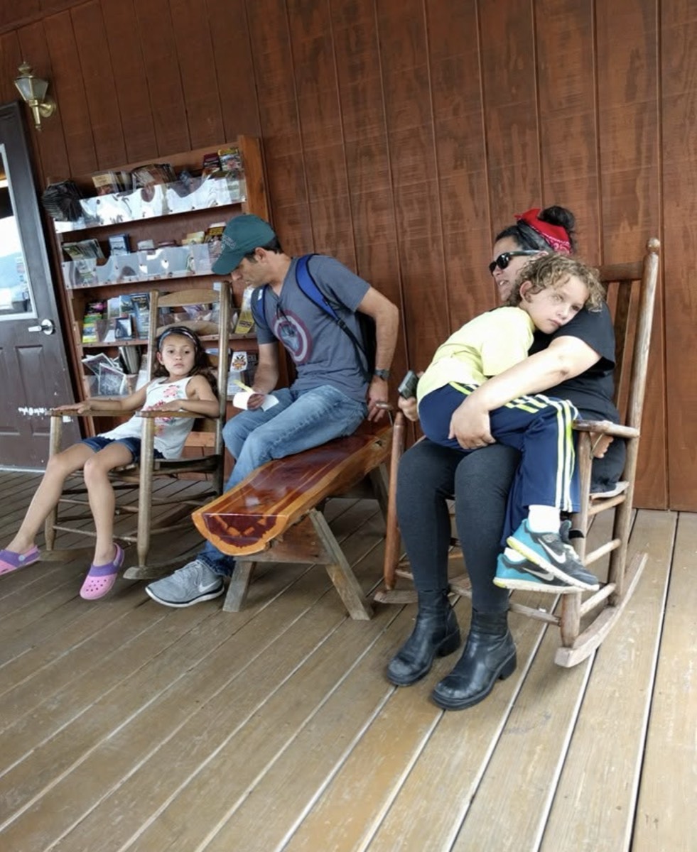 A girl in a rocking chair, a man in a ballcap on a bench and a woman with a boy asleep on her lap in another rocker, on a wooden porch beside a rack of postcards and guidebooks.