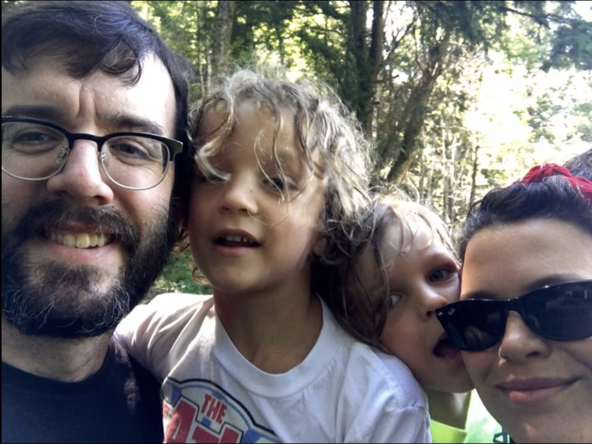 A close selfie of a bearded man in glasses, two children with windblown hair pulling faces, and a woman in sunglasses and a red headscarf, green woods behind them.