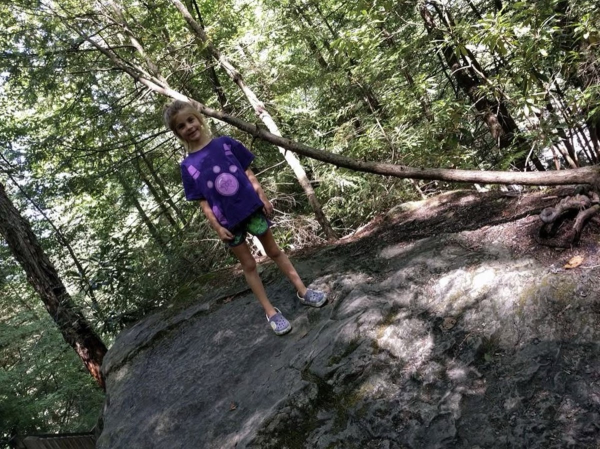 Brook standing on top of a large mossy boulder in the woods in a purple t-shirt and green patterned shorts, holding a low branch, smiling down at the camera.