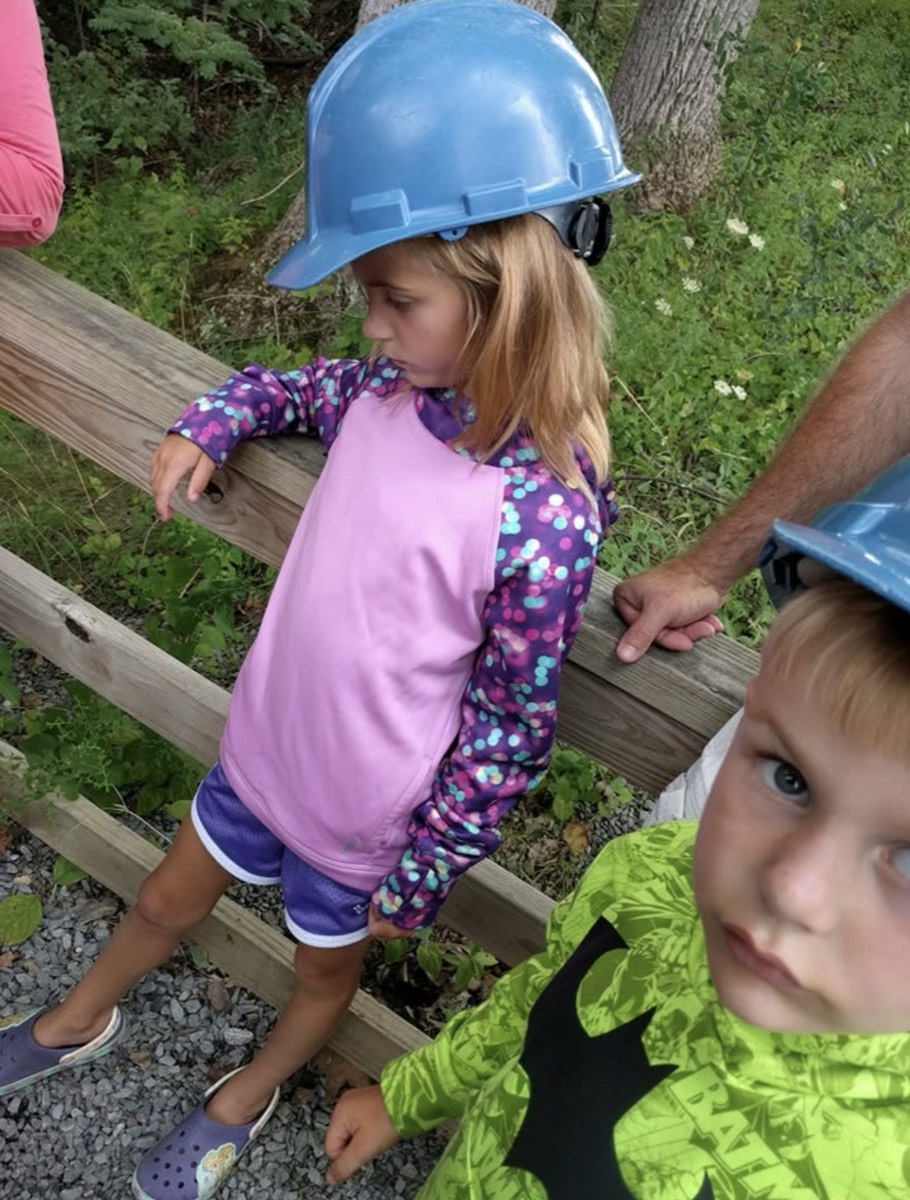 Two young children wearing oversized blue plastic hard hats issued for a cavern tour, standing beside a wooden rail fence and gravel path, an adult's hand resting on the rail behind them.