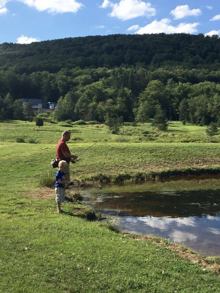 A man in a red shirt and a small boy in a blue shirt fishing side by side at the edge of a pond, mown grass and a forested ridge behind them under a bright blue sky.