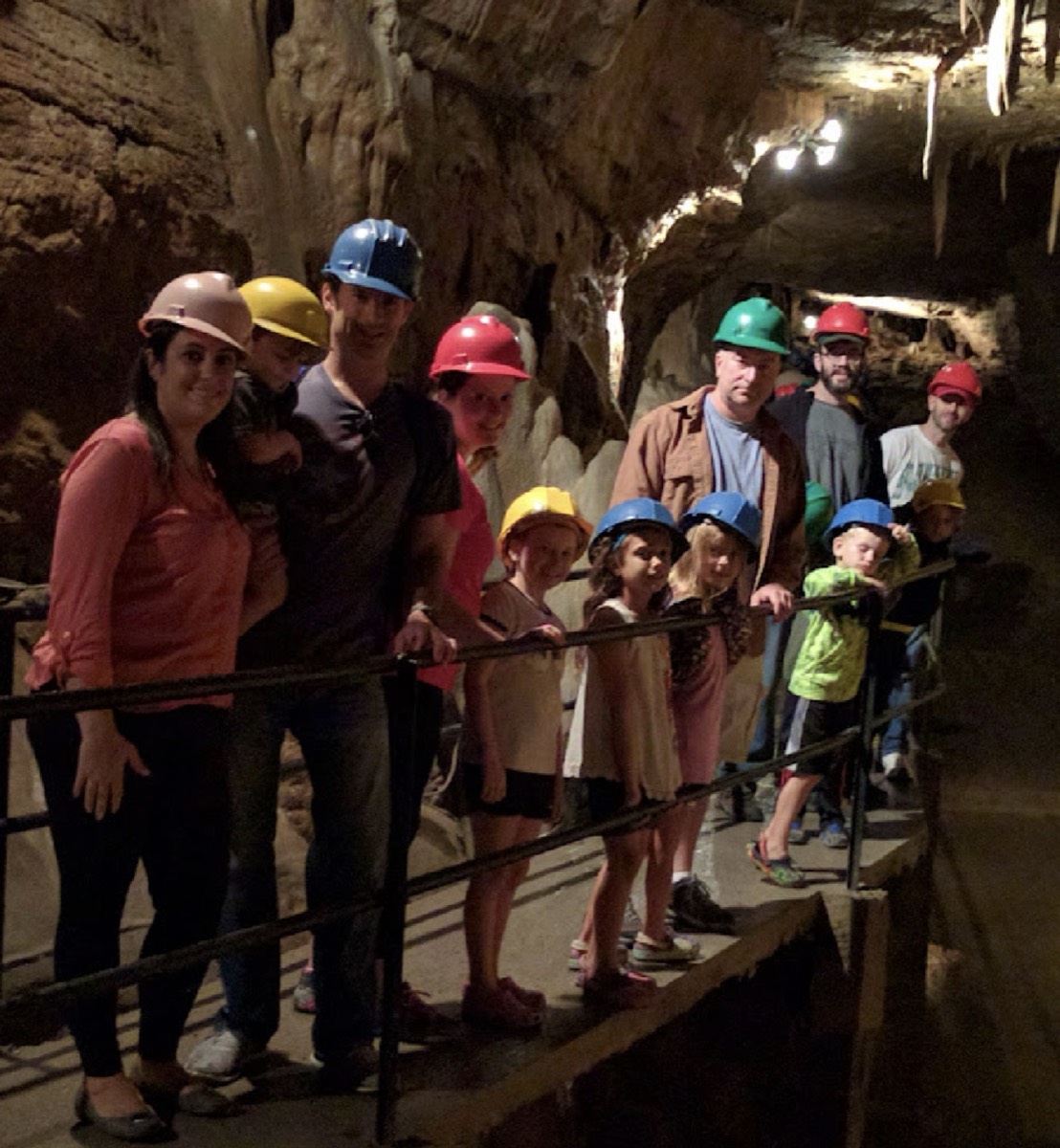 The gang lined up along a metal handrail on a walkway deep inside a limestone cavern, adults standing at the back and seven children in front, all wearing colored plastic hard hats, stalactites hanging from the ceiling above them.
