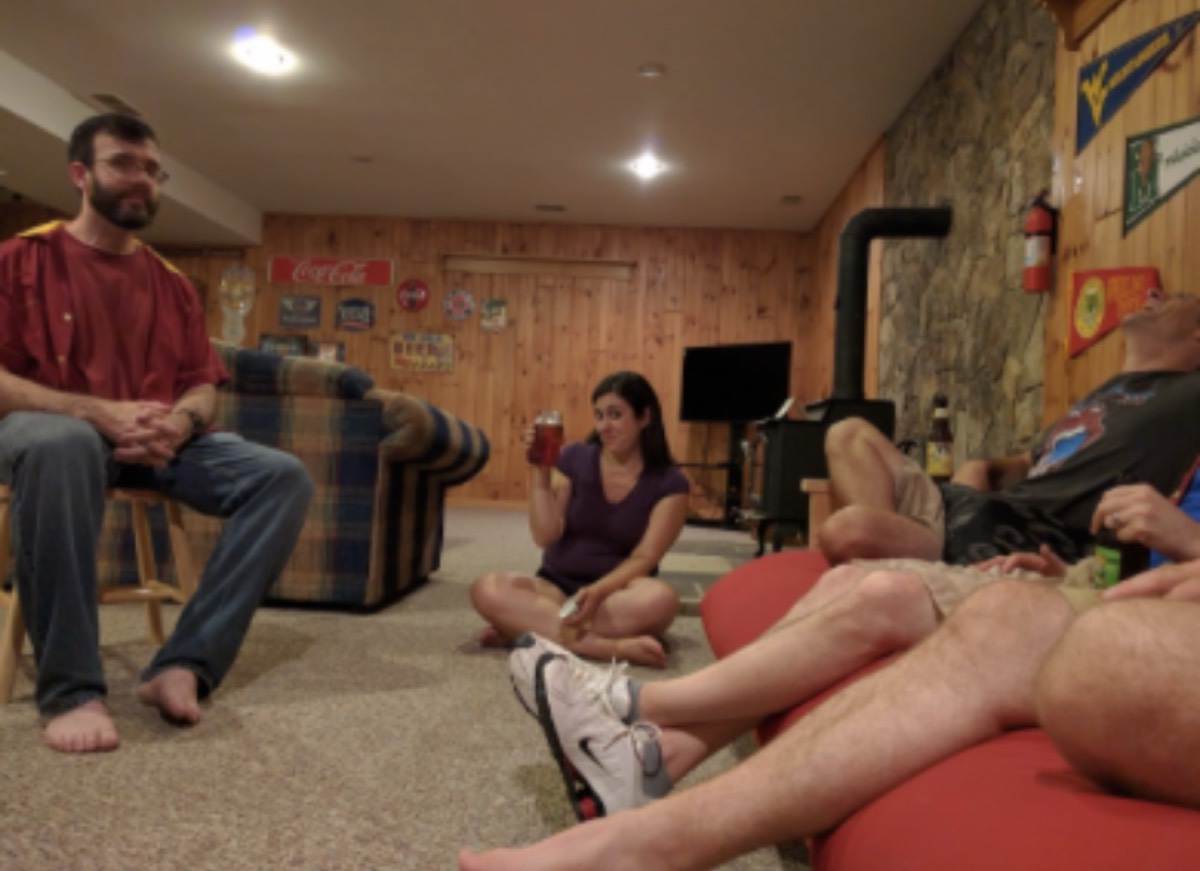 A wood-panelled basement rec room with a Coca-Cola sign and college pennants on the walls and a wood stove in the corner. A man sits in a wooden chair on the left, a woman sits cross-legged on the floor holding a drink, and others are stretched out on a red futon at the right of the frame.