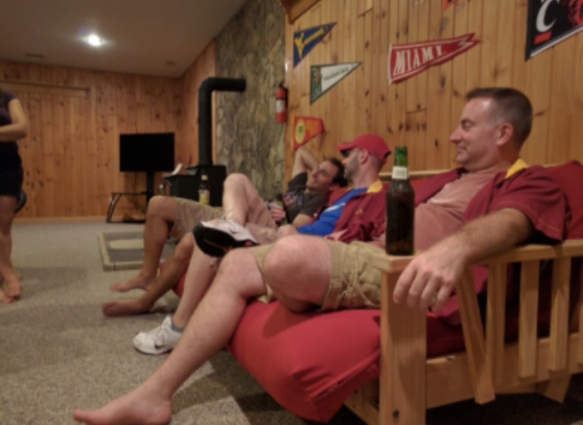 Three men reclining along a red futon in the wood-panelled basement, a beer bottle on the wooden arm rest beside them, college pennants for West Virginia, Miami and Cincinnati pinned to the wall above.