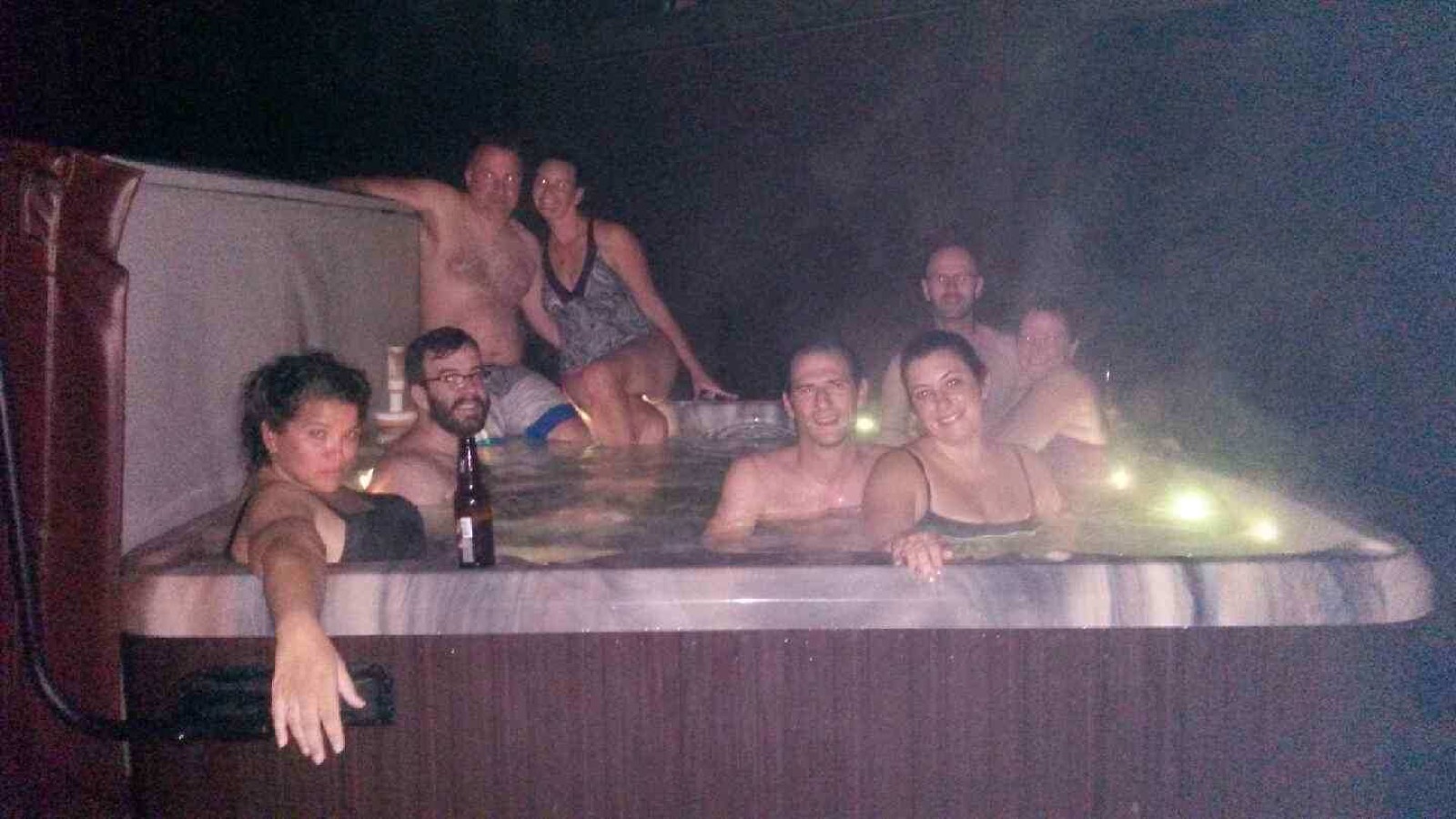 Eight adults crowded into a steaming hot tub at night, beers in hand, lit only by the tub's underwater lights.