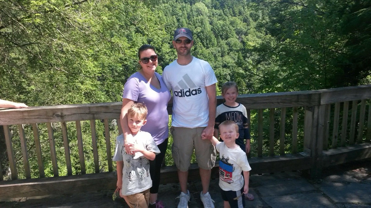 The Nardella family of five at the Blackwater Falls overlook railing. Left to right: Ryan in a grey bug-print tee, Kelly in a lavender v-neck and sunglasses, Shawn in a white Adidas tee and ballcap, Owen in a white fire-truck tee, and Claire in a navy MEOW tee, the wooded gorge dropping away behind them.