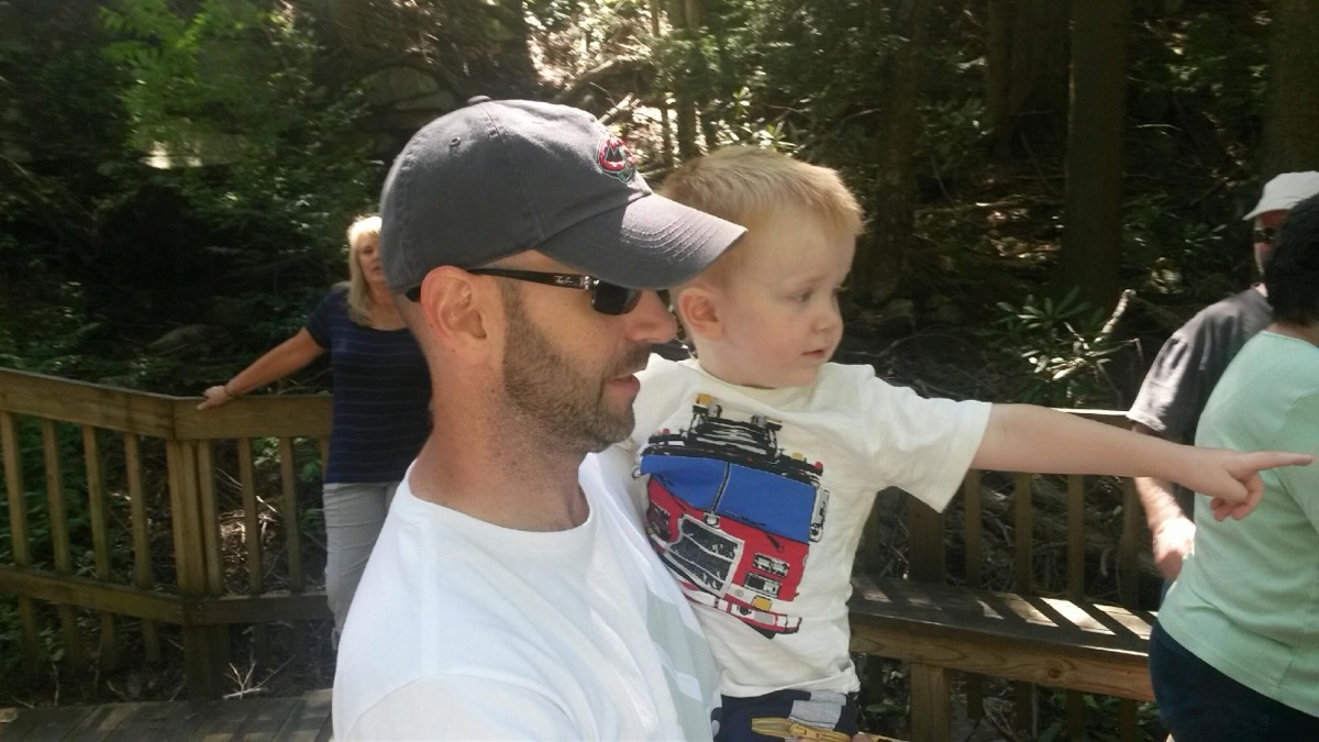A dad in a Cleveland ballcap carrying a toddler in a fire-truck shirt who is pointing off into the trees along a boardwalk.