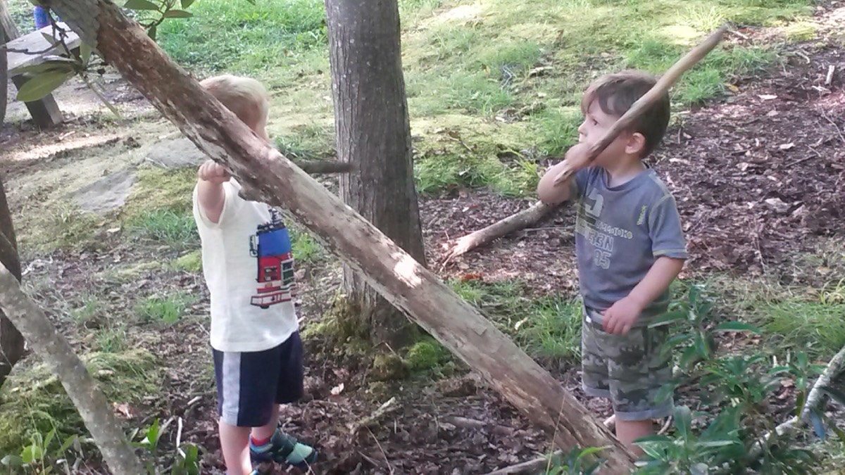Two boys in a wooded cabin yard, each holding a long stick like a sword.