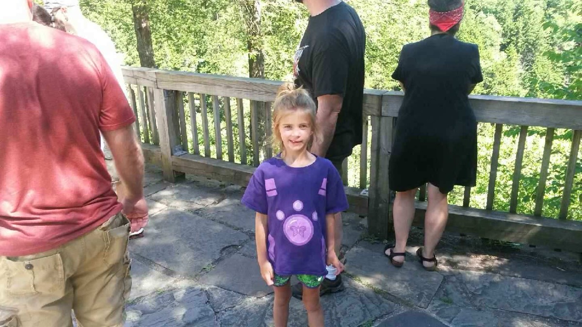 A young girl in a purple shirt with a round medallion standing on the stone overlook platform, adults at the railing behind her.