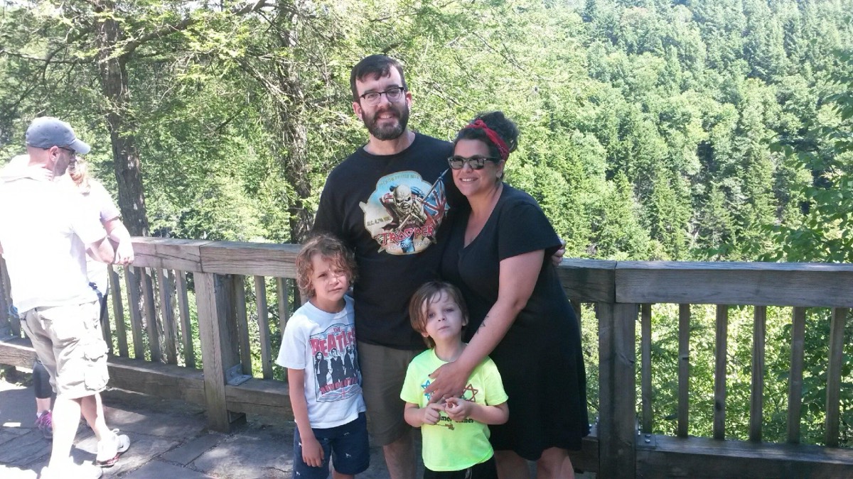 A family of four posing at a wooden overlook railing with a deep green forested gorge behind them.