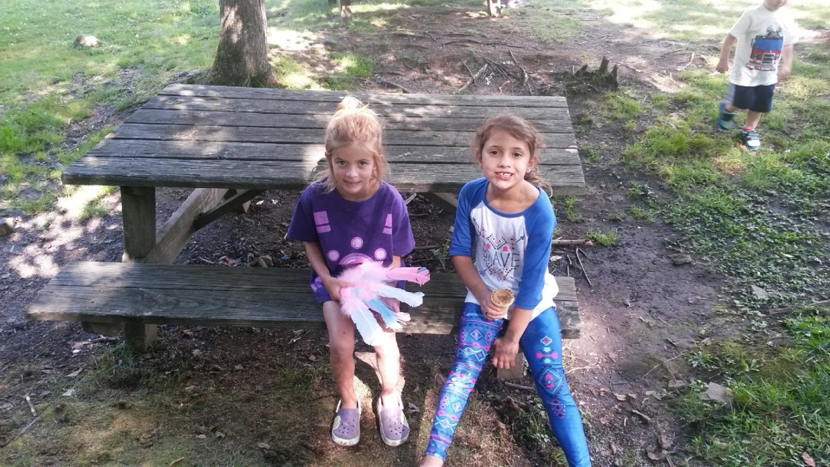 Two young girls sitting on a weathered wooden picnic table in the cabin yard, one holding a pink toy glove.
