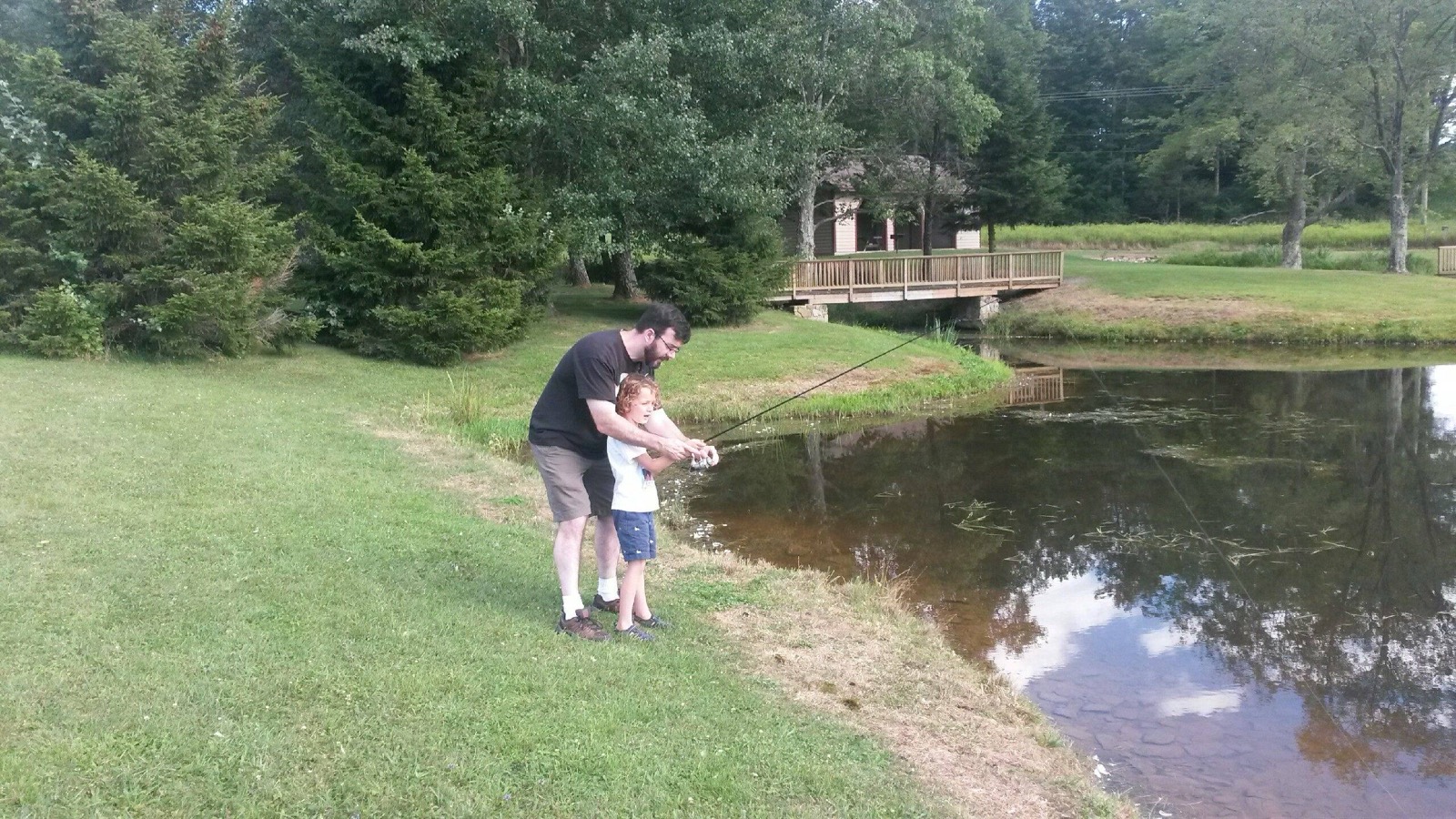 A dad helping a small child cast a fishing rod at the edge of a still cabin pond, a cabin with a deck on the far bank.