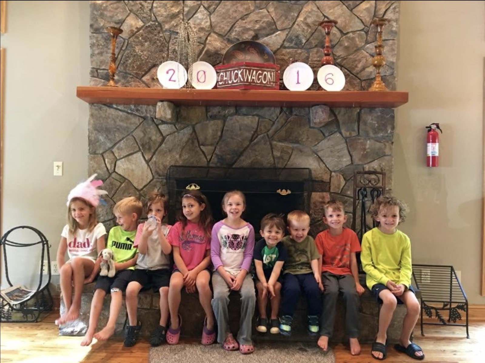 Nine children sitting in a row on a stone fireplace hearth, the mantel above them decorated with paper plates spelling 2016 and a CHUCK WAGON sign.