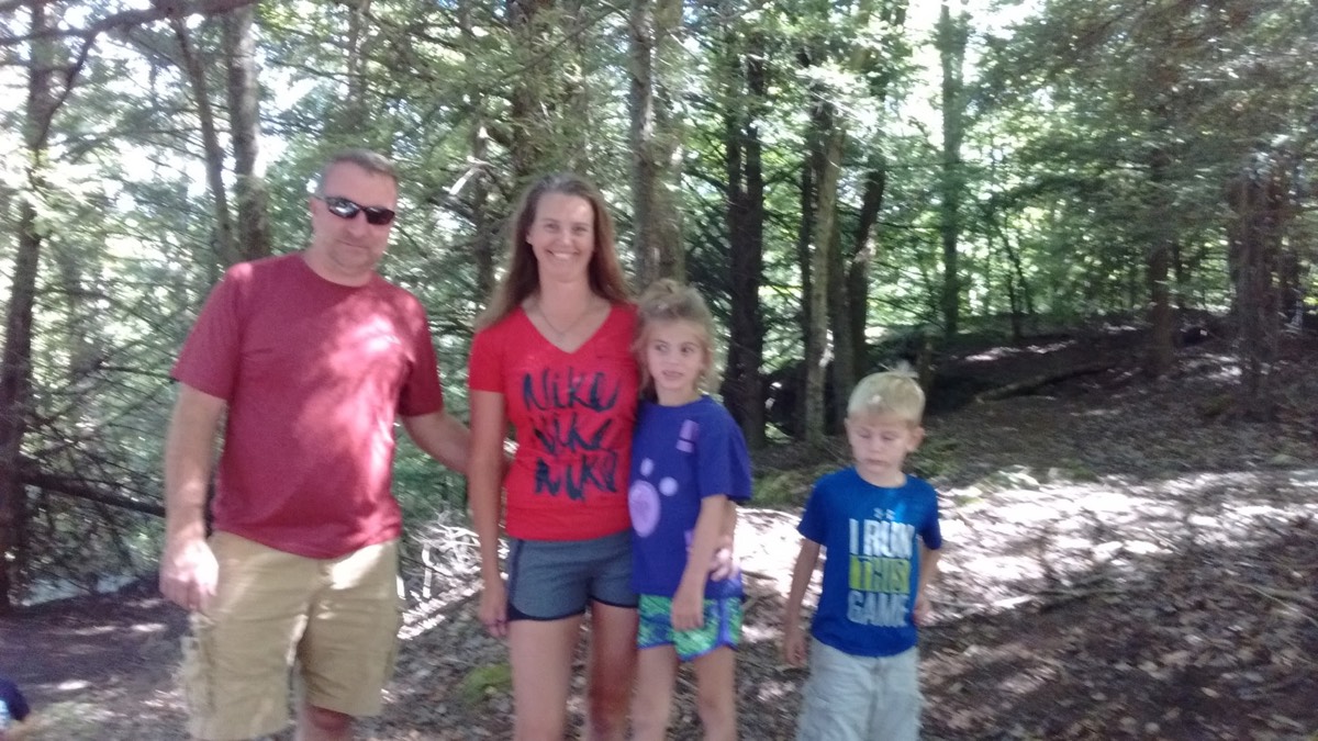 The Lockard family of four posing on a wooded trail: Heath in a maroon shirt and sunglasses, Mandy in a red Nike shirt, Brook in a blue shirt, and Colton in a blue shirt.