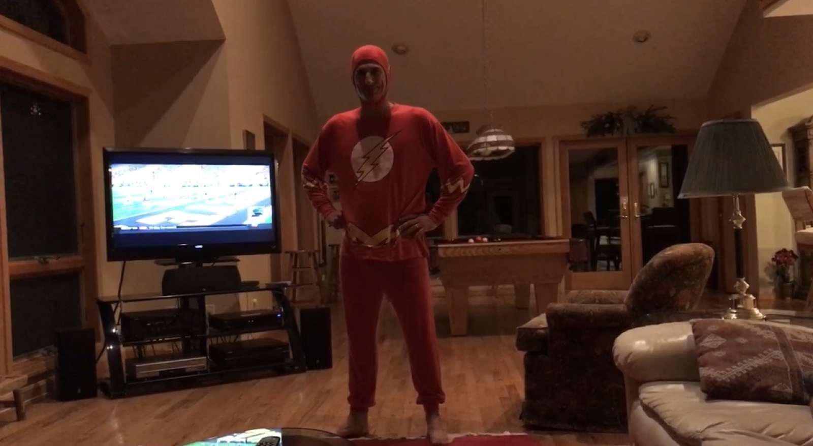 A grown man standing hands-on-hips in a cabin living room wearing a full red Flash costume with hood and lightning-bolt emblem, a football game on the TV and a pool table behind him.