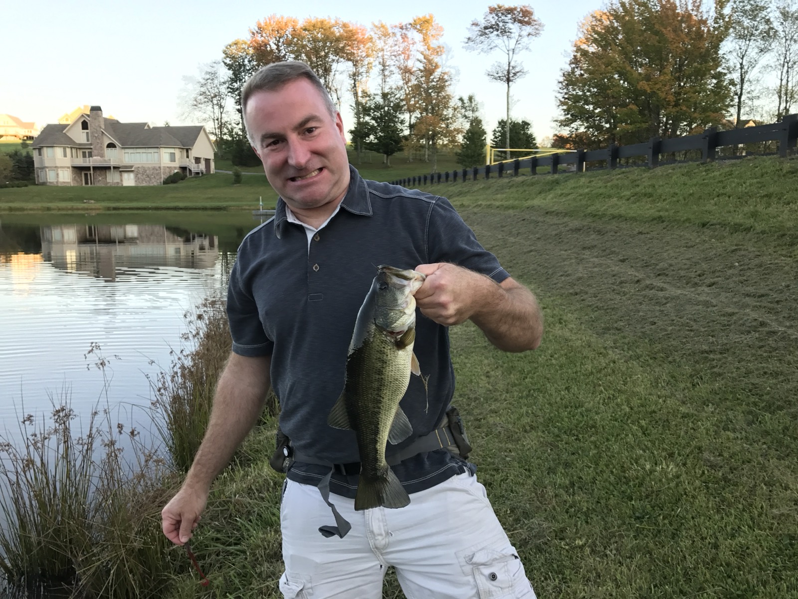 Heath holding up a largemouth bass by the mouth at the edge of a pond at sunset, a large house and turning fall trees across the water behind him.
