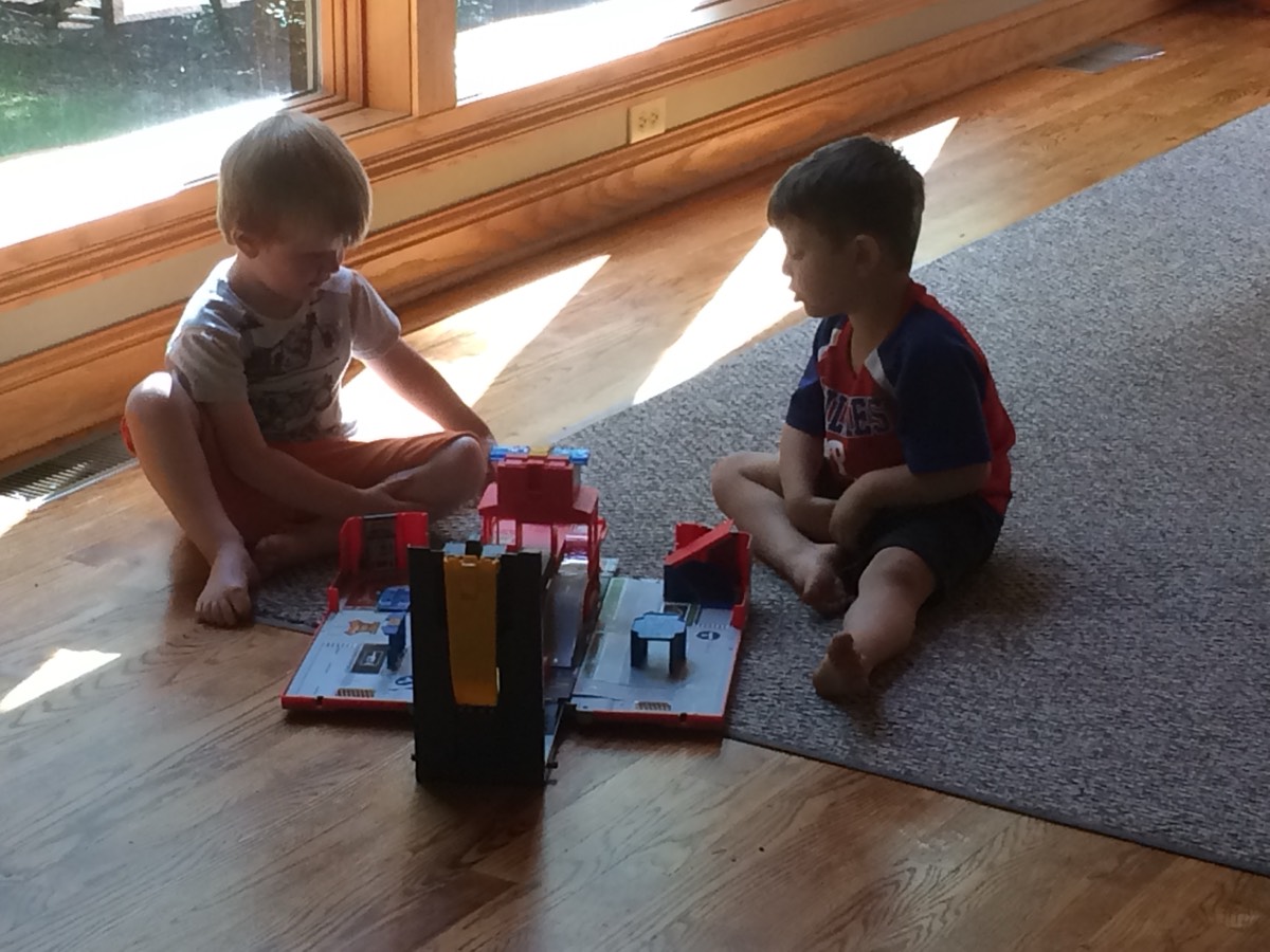 Two young boys sitting cross-legged on the floor by a wall of windows, playing with a red toy fire-station playset between them.