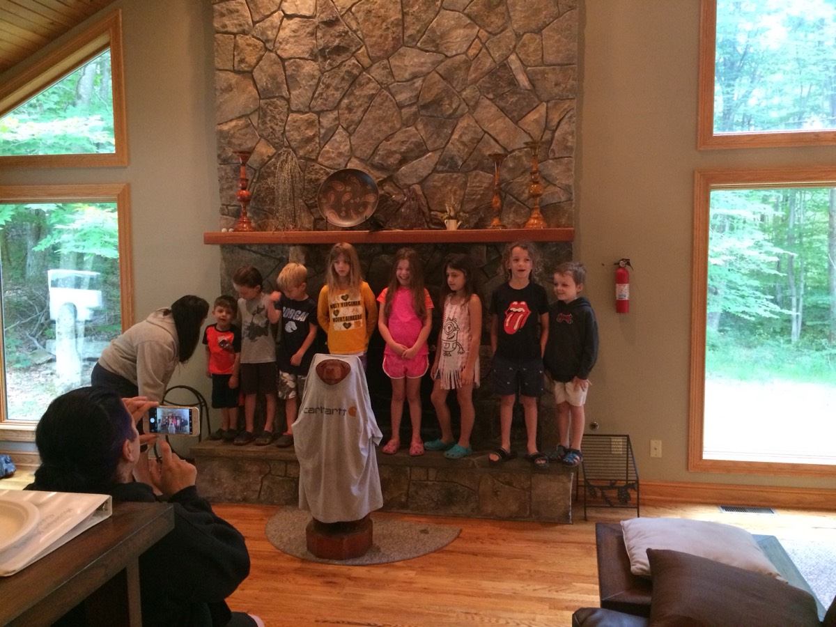 A woman leaning in to photograph a row of children on a stone hearth with her phone, a carved bear statue draped in a Carhartt shirt in the foreground.