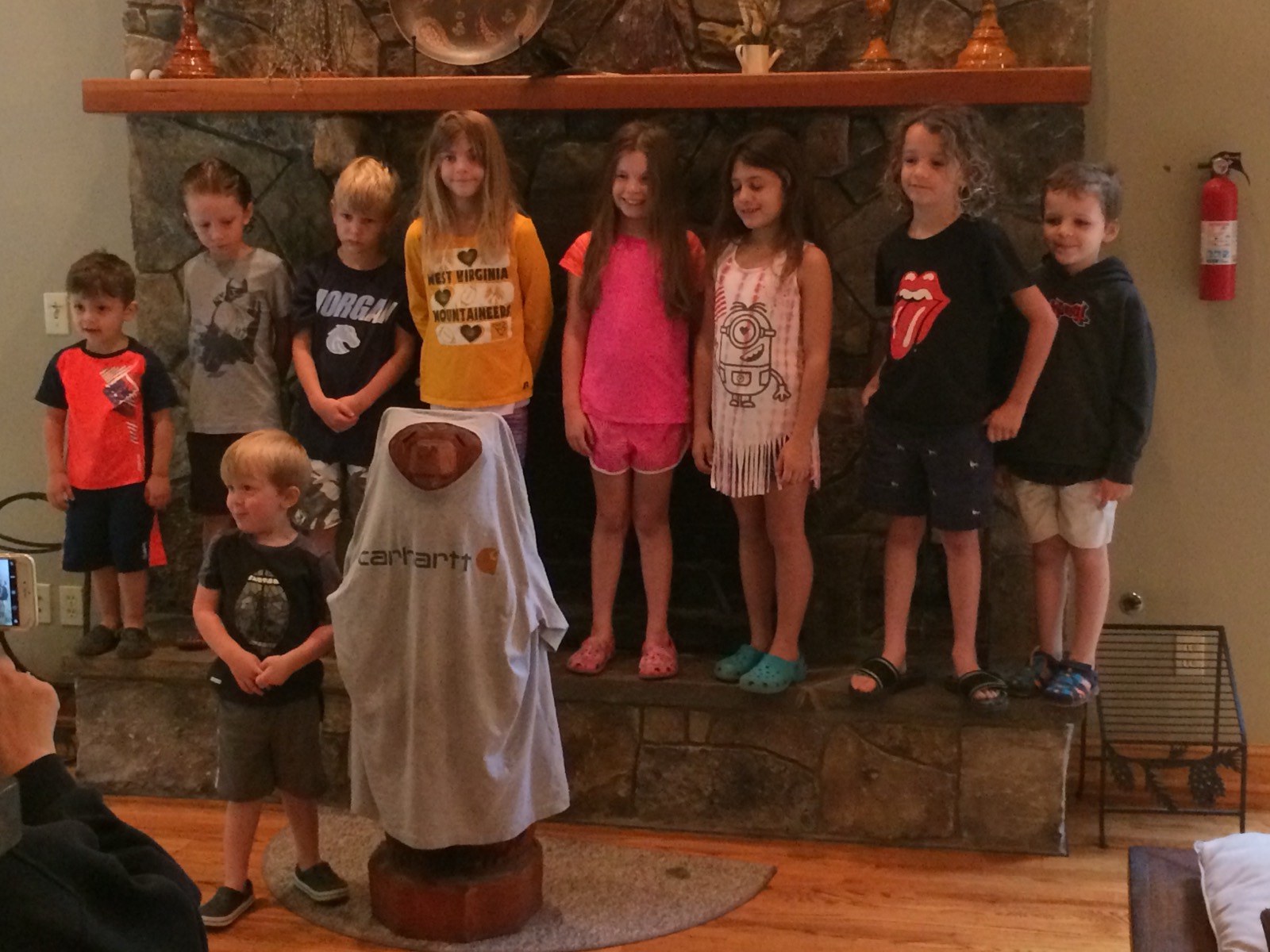 Nine children posed in a row on a stone fireplace hearth, a carved wooden bear in a Carhartt shirt standing in front of them.