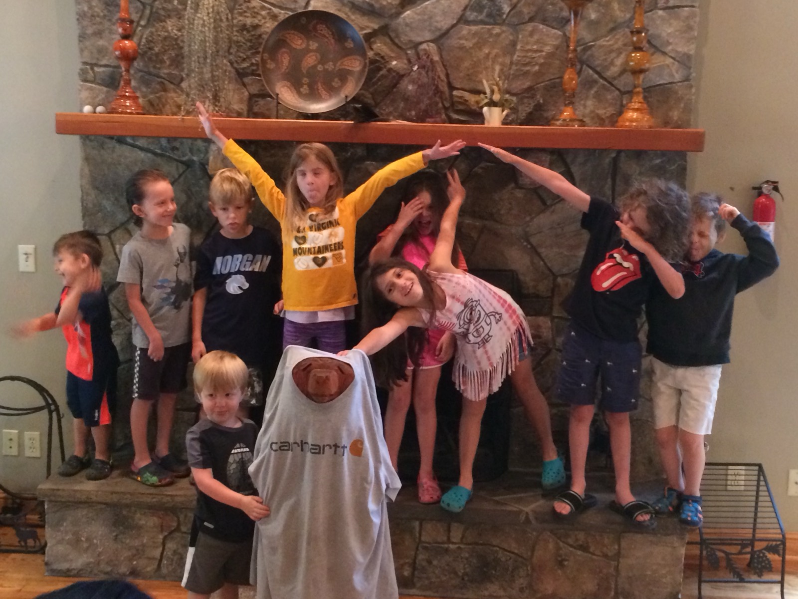 The same nine children on the hearth now goofing off, arms thrown up, tongues out, one covering her eyes.