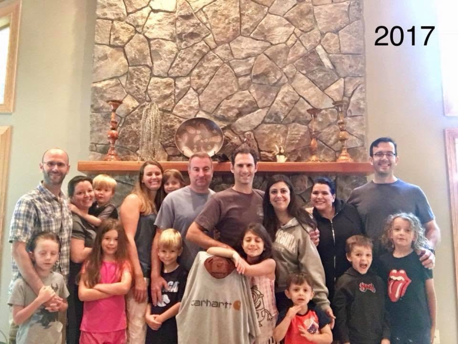 The full Chuckwagon Gang posed in front of a tall stone fireplace, adults standing behind a row of children, a carved wooden bear in an oversized Carhartt shirt at the center front; the year 2017 is written in the top right of the photo.