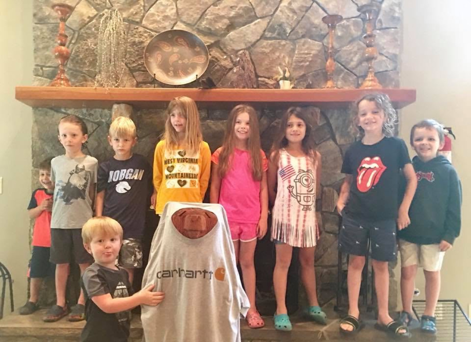 Nine children standing in a row along the stone hearth ledge in front of the fireplace, with a carved wooden bear wearing an oversized tan Carhartt shirt on the floor in front of them.