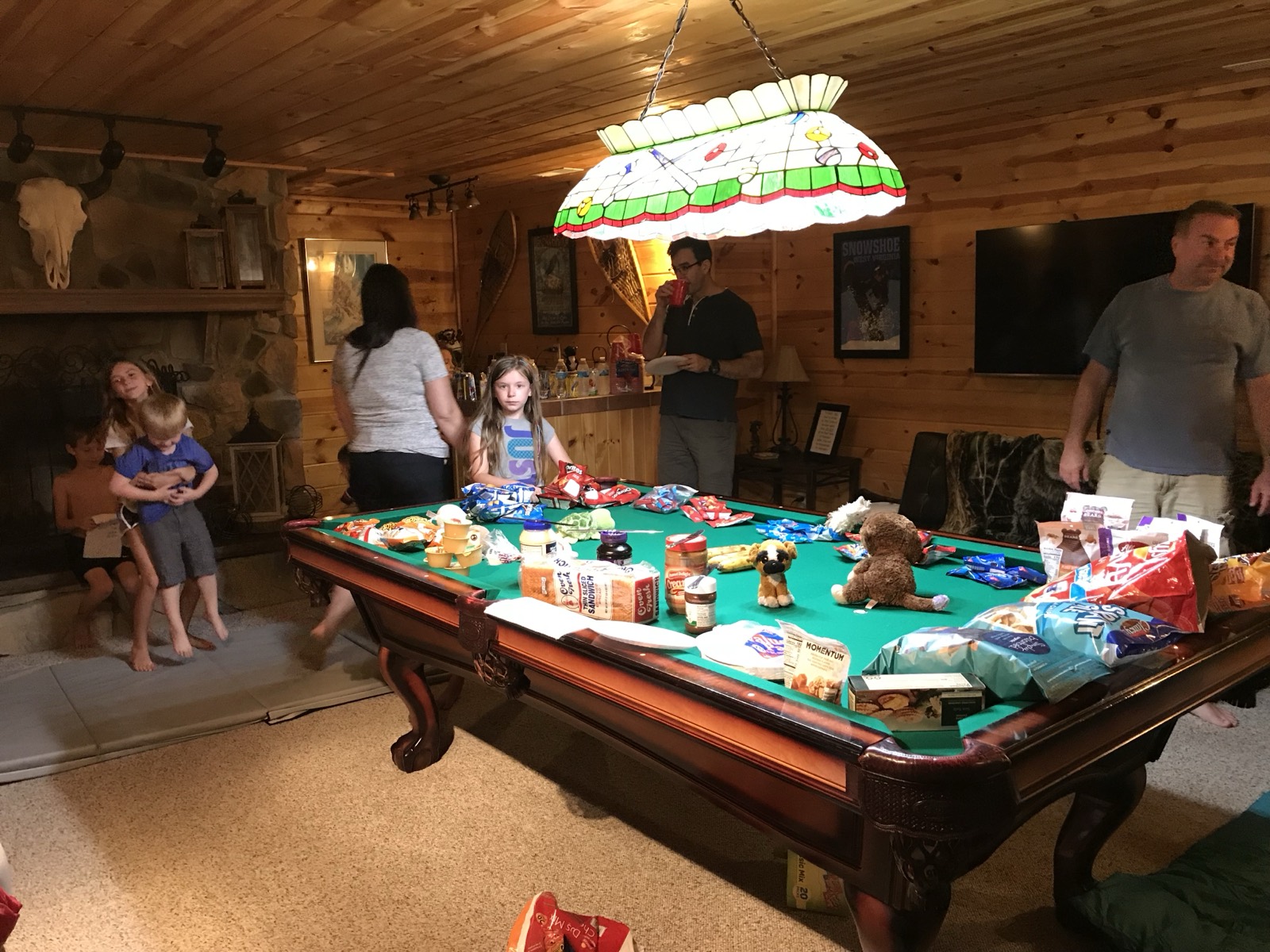 A cabin game room where a pool table is completely covered in bread, snacks, chip bags and stuffed animals, kids and adults around the edges, a stained-glass lamp overhead and a Snowshoe West Virginia poster on the wall.