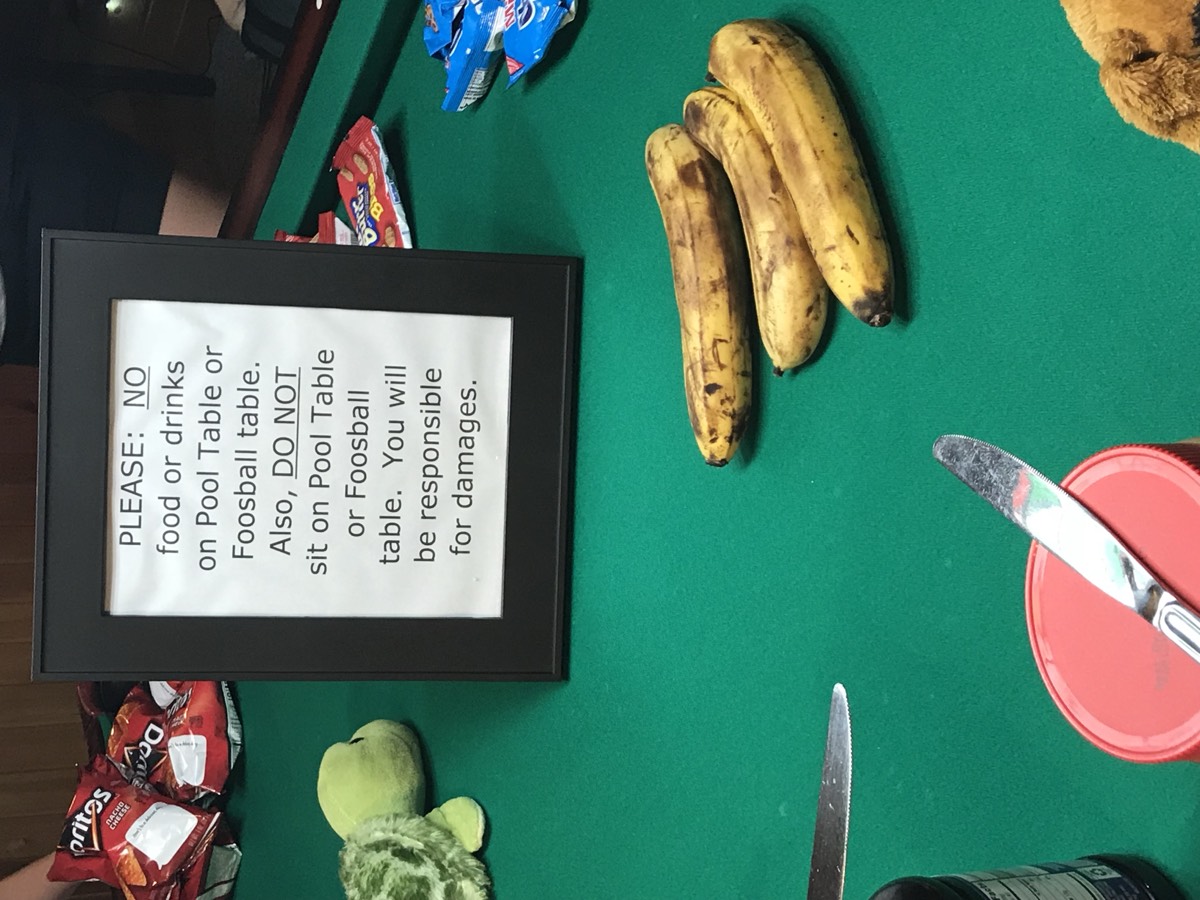 A framed sign reading PLEASE: NO food or drinks on Pool Table or Foosball table, sitting on green pool-table felt surrounded by bananas, Doritos, a knife and snack wrappers.