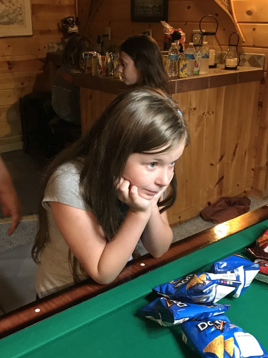 A girl leaning on the rail of a pool table with her chin resting in her hands, open bags of Doritos on the green felt in front of her.