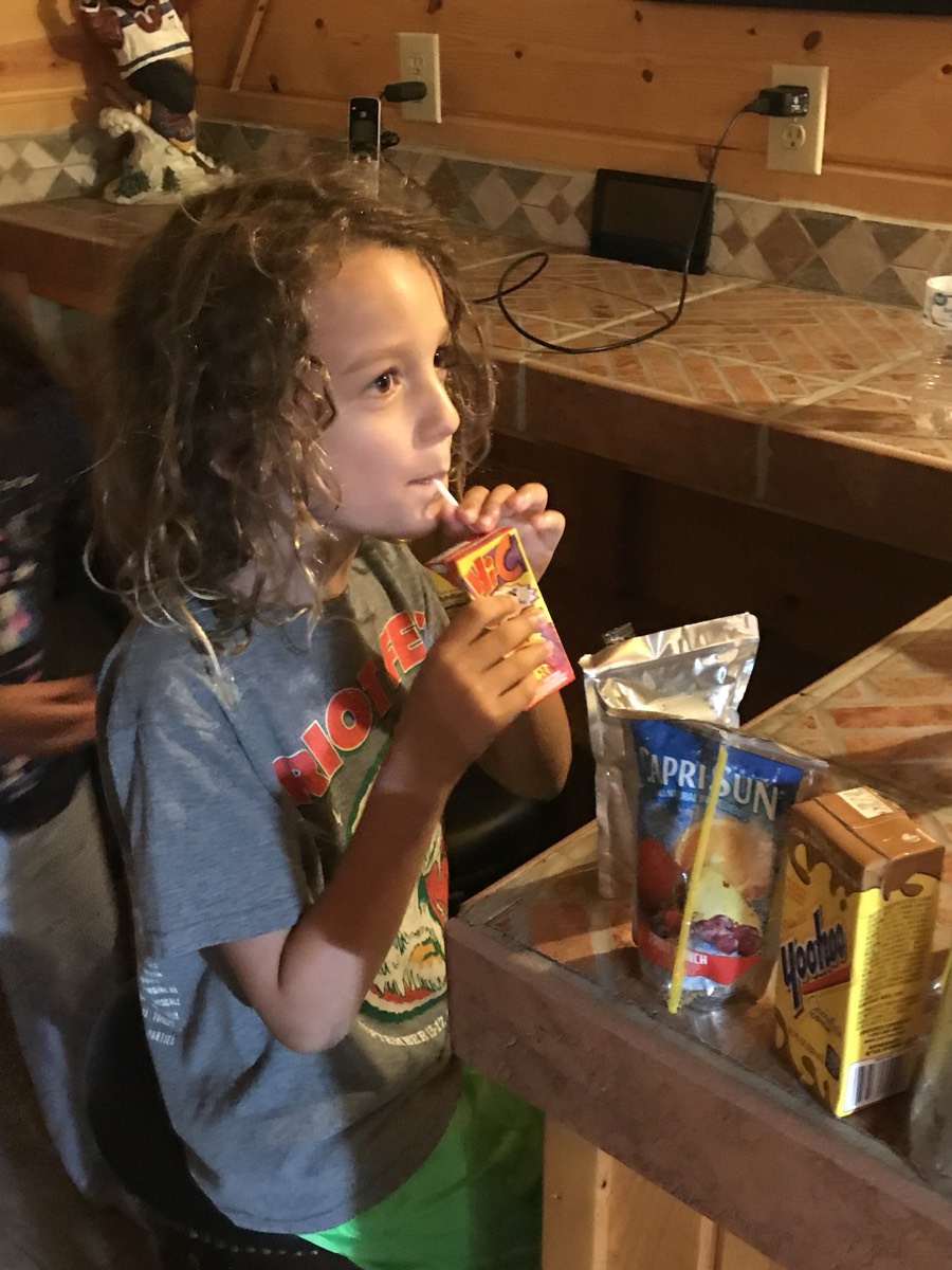 A curly-haired child in a gray Riot Fest t-shirt sipping a Hi-C juice box through a straw, a Capri Sun and Yoo-hoo on the counter beside him.