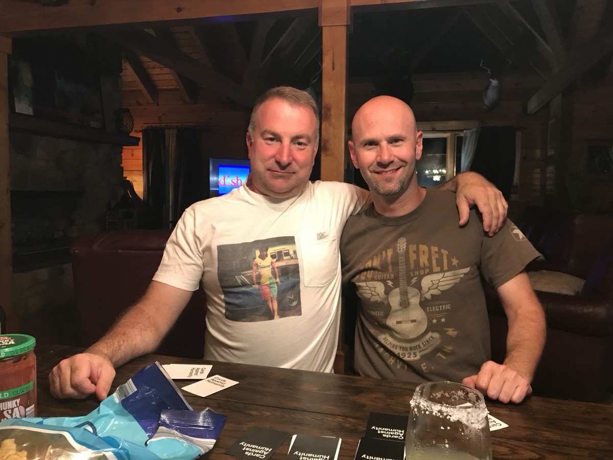 Two grown men arm over shoulder at a cabin table at night; the man on the left wears a white t-shirt printed with a photo of a teenage boy in a cut-off shirt and colorful jams shorts with a mullet, posing by a car. Cards Against Humanity cards and a salt-rimmed margarita on the table.