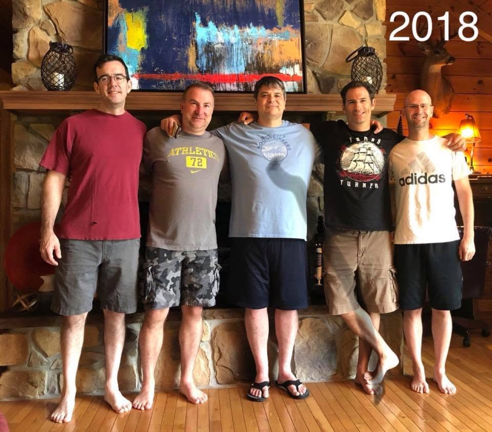 Five men standing in a row in front of the cabin's stone fireplace, a large abstract painting on the mantel above them; the year 2018 is written in the top corner of the photo.