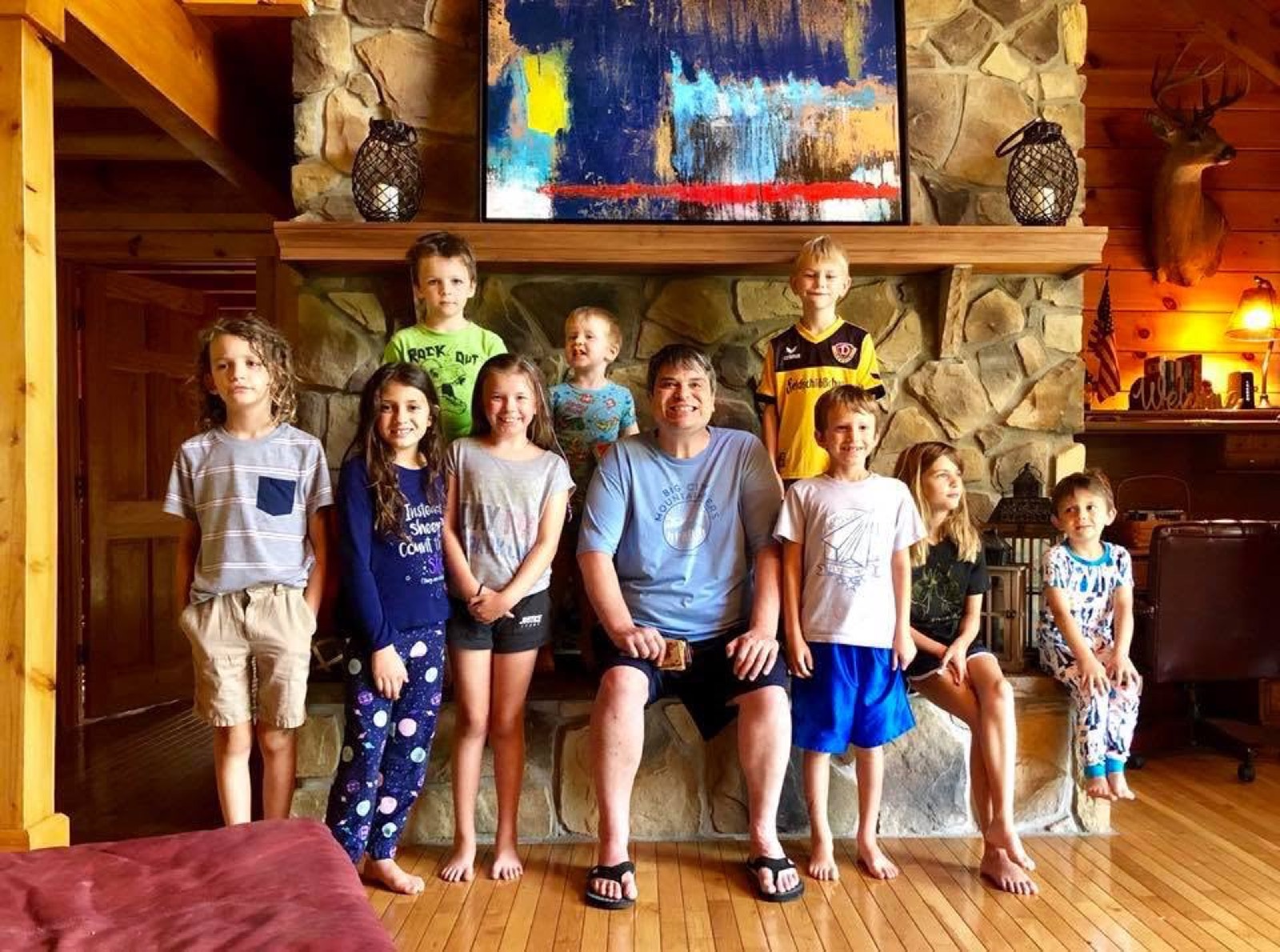 Nine children arranged on and around a stone fireplace hearth in pajamas and shorts, an abstract painting and two lanterns on the mantel behind them.